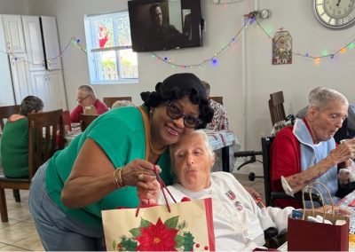 Smiling caregiver in green top leaning affectionately next to a male resident holding a Christmas gift bag in a festively decorated dining room at Paradise Manor assisted living facility in Florida