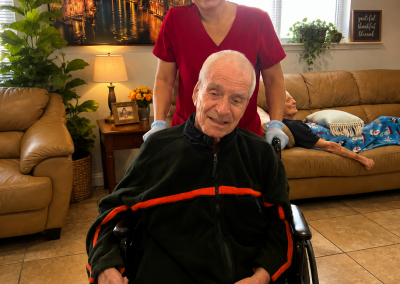 Female caregiver in red scrubs assisting an elderly male resident in a wheelchair in the living room of a Florida ALF