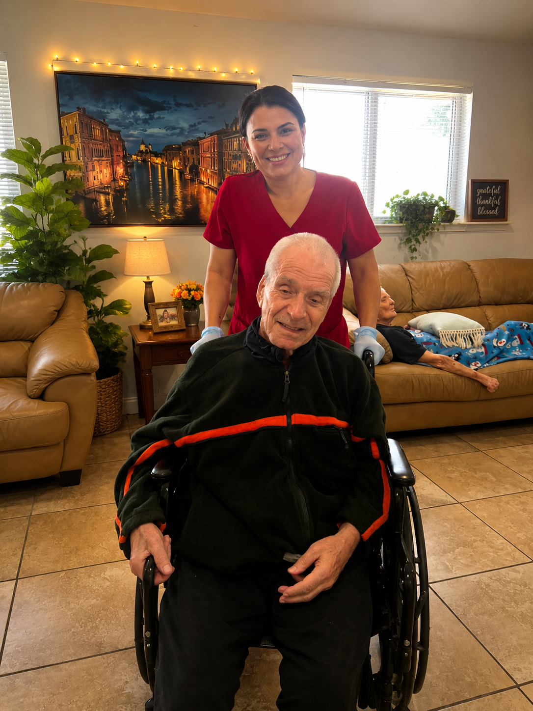 Female caregiver in red scrubs assisting an elderly male resident in a wheelchair in the living room of a Florida ALF