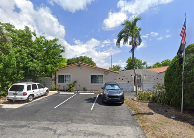 Exterior front view of Paradise Manor senior assisted living facility in Florida, a beige stucco building with a red tile roof, palm tree, American flag, lush greenery, and private parking lot on a sunny day