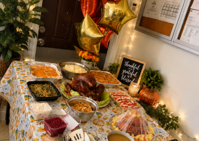 Thanksgiving holiday feast buffet table with roasted turkey, mashed potatoes, green beans, stuffing, gravy, cranberry sauce, and fresh fruit at Paradise Manor male-only assisted living facility in Florida, decorated with gold and orange star balloons, string lights, pumpkins, and a "Thankful Grateful Blessed" chalkboard sign